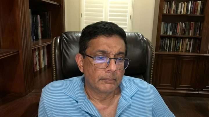 Man wearing blue striped polo and glasses sits in a leather chair in a home library, looking at the camera for a neutral expression.