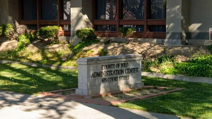 Stone sign reads 'County of Yolo Administration Center, 625 Court Street' in front of a government building with landscaping and brick facade rear windows.
