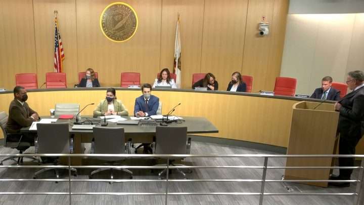 A formal meeting in a council chamber with people seated at a long table and others at a raised panel, microphones and documents in use.