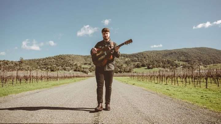 Man standing in the middle of a gravel road, playing an acoustic guitar, with vineyard rows and hills in the background.