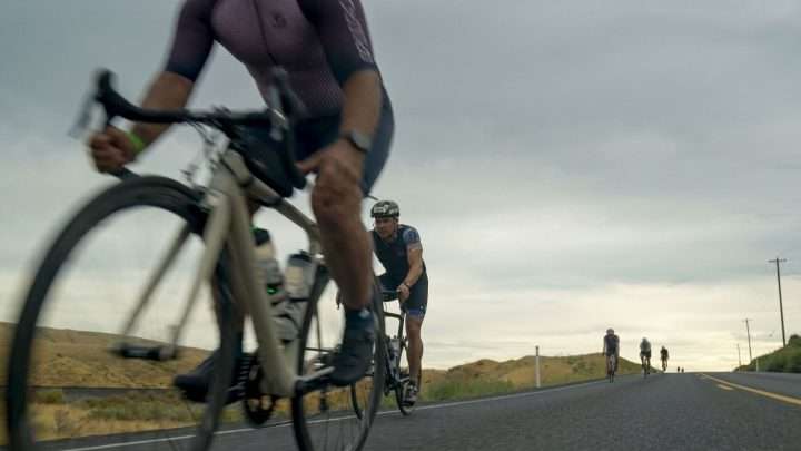 Cyclist in foreground rides a road bike on a rural highway as others pedal in the distance under a cloudy sky.